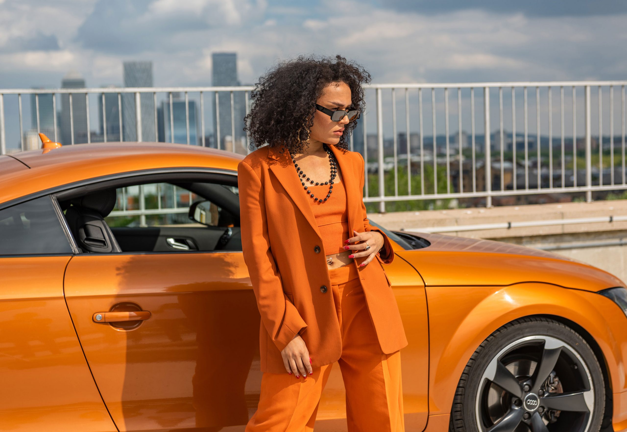 A fashion model wears an orange suit and sunglasses, posing in front of an orange sports Audi TT car. Shot for an ad campaign for the social car hiring platform Turo, this image was captured on a rooftop multistorey car park overlooking London.