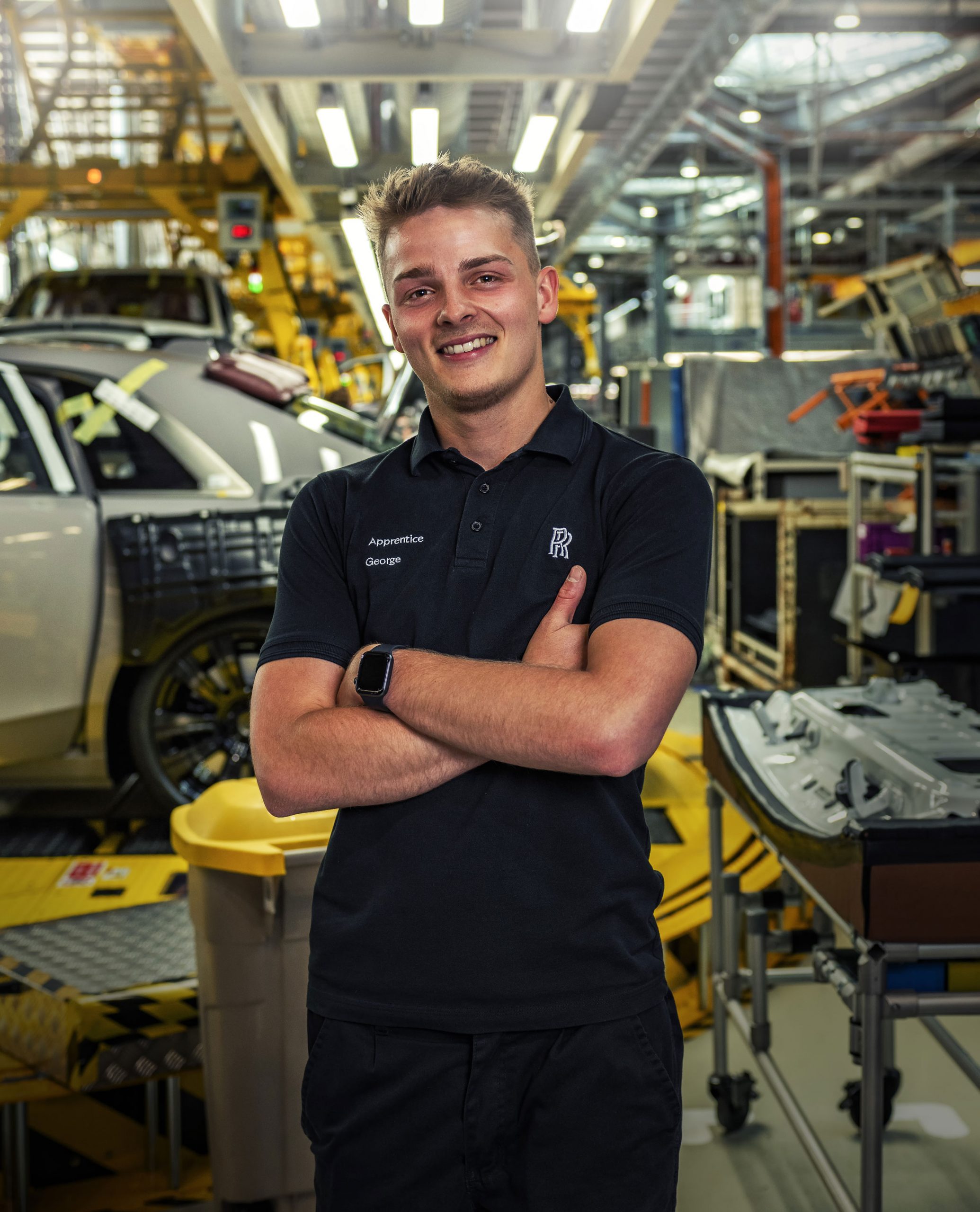 Young male apprentice from Rolls-Royce Motor Cars exudes confidence and pride while working on the production line. Folding his arms and with a happy expression on his face, he stands in front of the luxury vehicles he helps produce, showcasing the skilled craftsmanship of the company.
