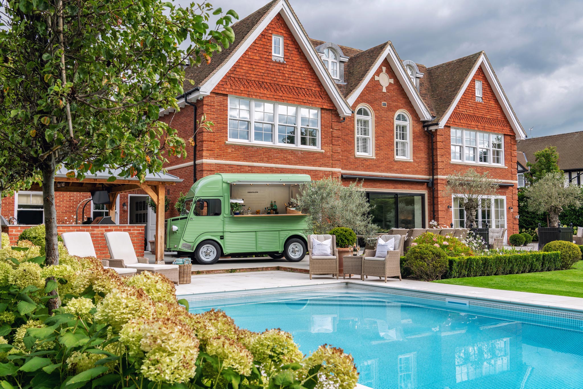 Vintage van parked next to a sparkling swimming pool surrounded by a lush garden. The beautiful home in the background adds a touch of luxury to the idyllic outdoor setting. Perfect for a summer getaway or a staycation in style.