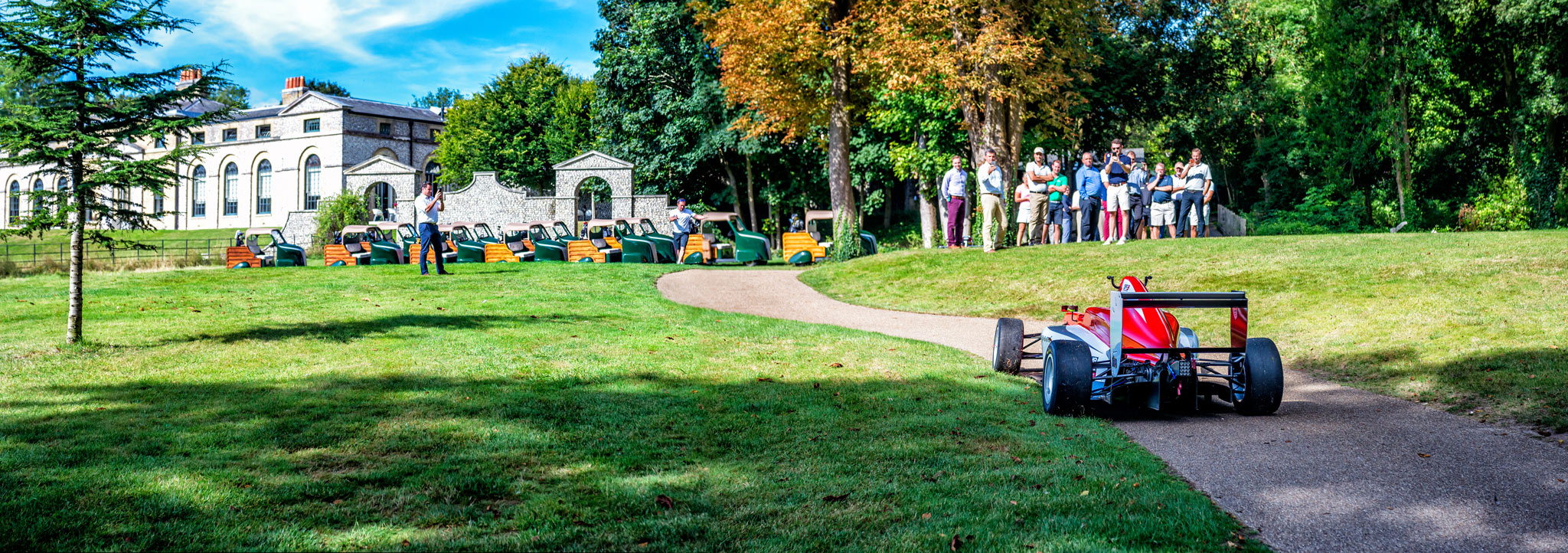 A scene of ambition captured by Wright Content, featuring a formula car driving up the fairway of a golf course. This promotional event aimed to showcase a young driver and attract potential sponsors, incorporating a day of golf for attendees. The image includes a crowd of people watching the car drive up the grass, with a majestic Manor House in the background. Wright Content specializes in capturing the spirit of athletic ambition and promotional events.