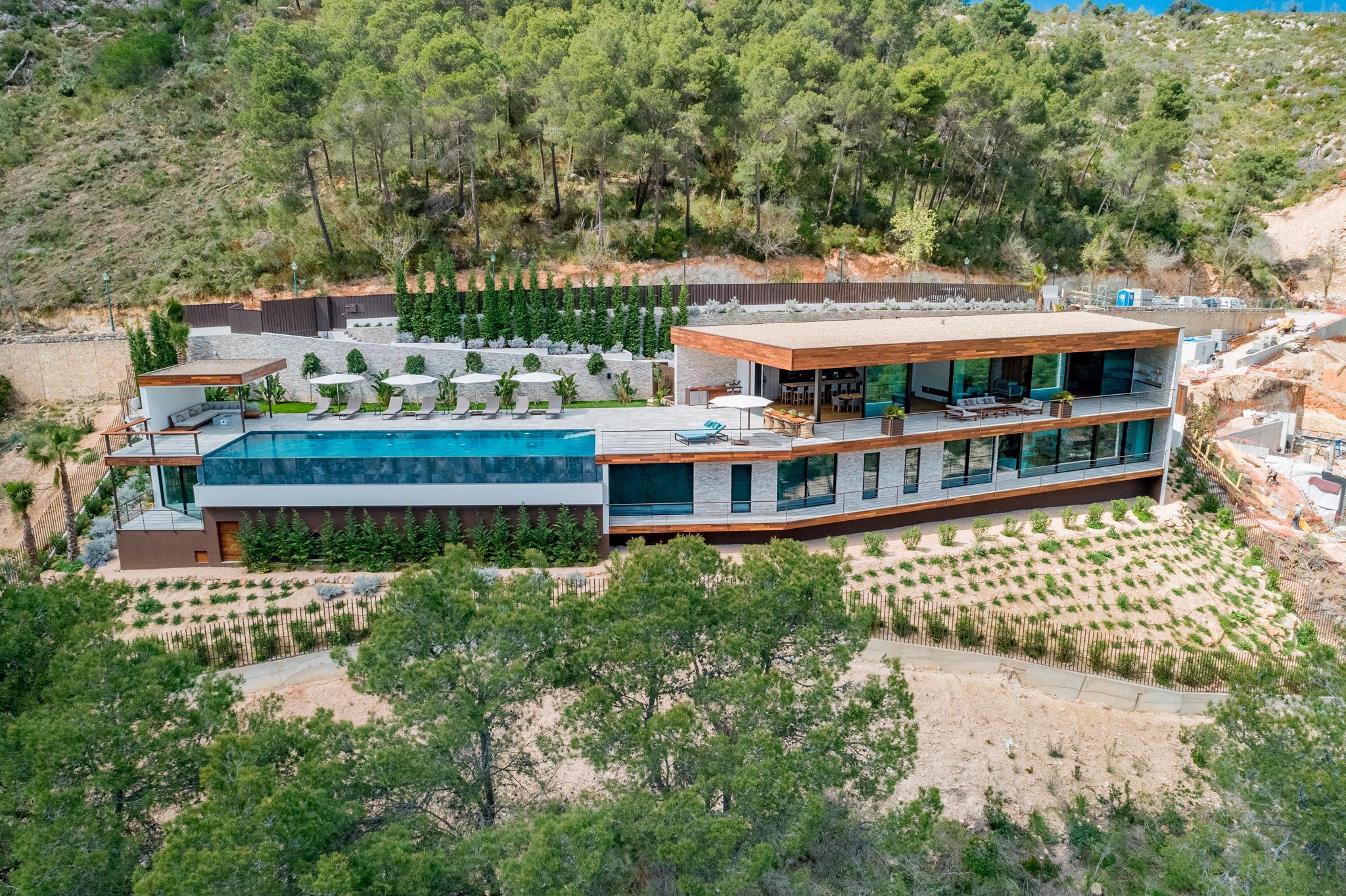 Aerial view of a modernist villa nestled in the Son Vida hills of Palma, captured by Wright Content. The luxurious home features multiple glass-fronted rooms, a spacious patio, and a pool, surrounded by lush greenery.