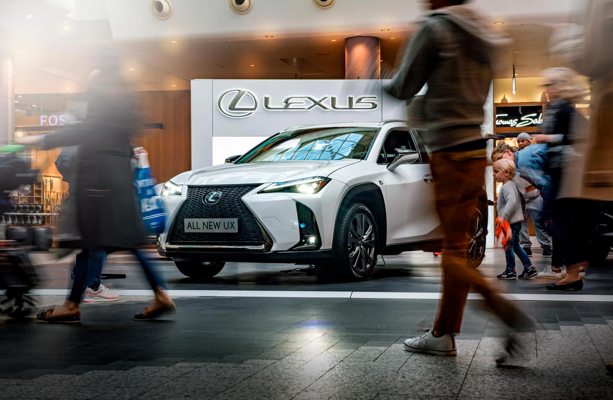 A captivating slow shutter speed image captures the bustling activity at a Lexus stand in a busy high-end shopping center in London. The focal point is a pristine white Lexus car, surrounded by people exploring the exhibit.