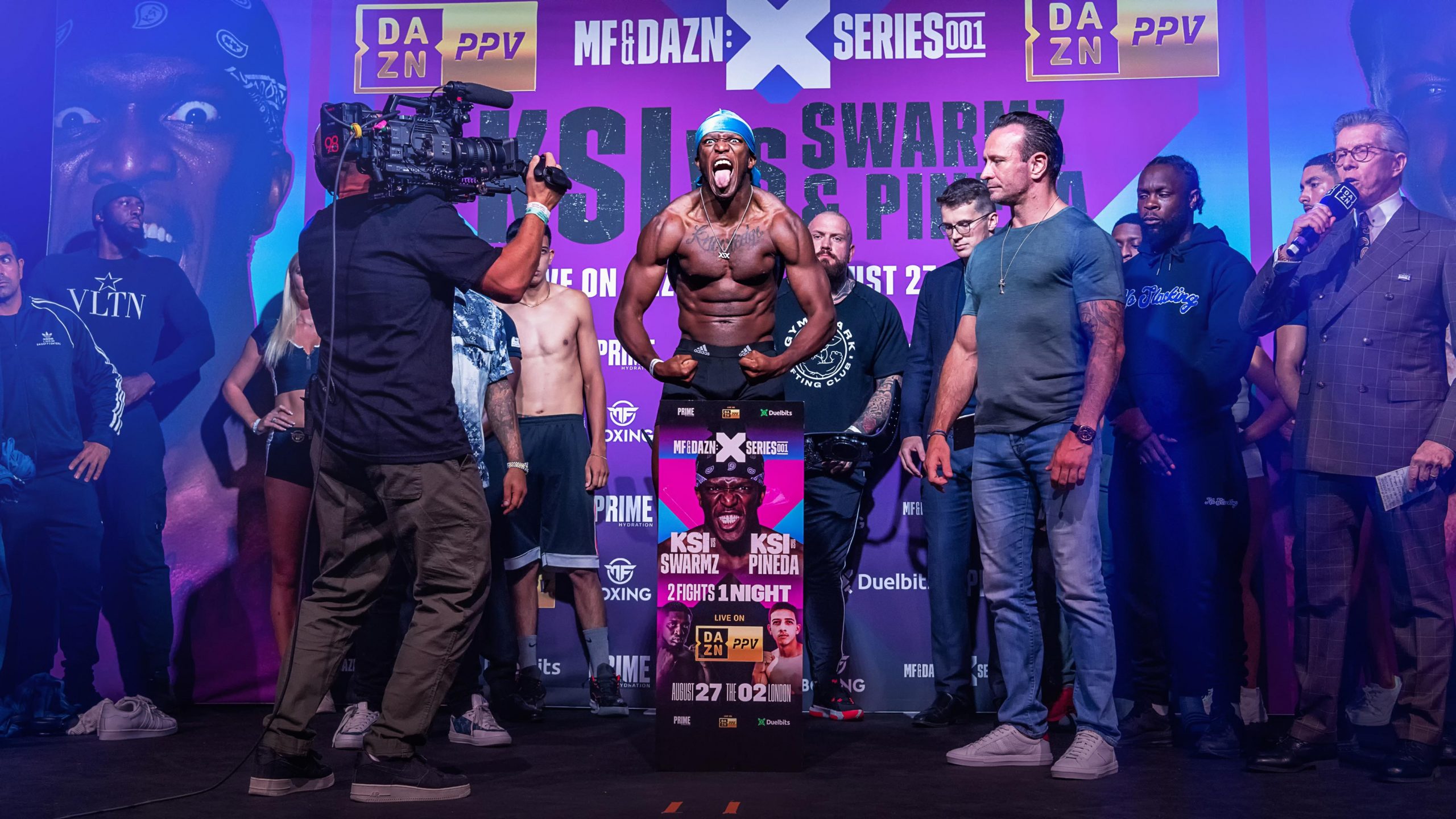 KSI flexes his muscles and sticks his tongue out, roaring, at a weigh-in for a large boxing match. The stage is filled with promoters, other fighters, and camera crews.