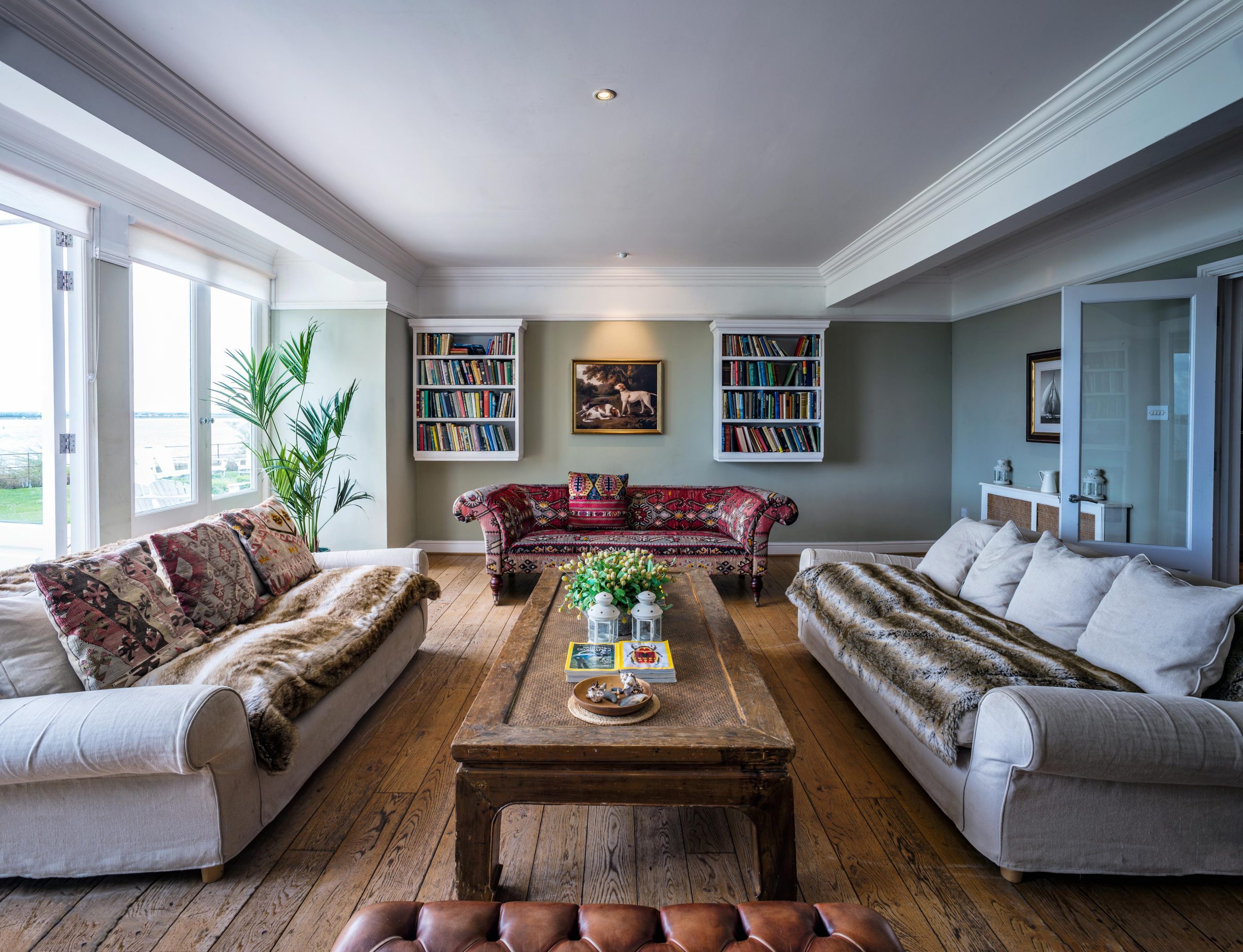 Inviting image of a cottage's spacious living room captured by Wright Content for an Airbnb rental on the outskirts of London. Large comfortable sofas, bookshelves, and a window overlooking a private beach create a cozy and relaxing atmosphere.