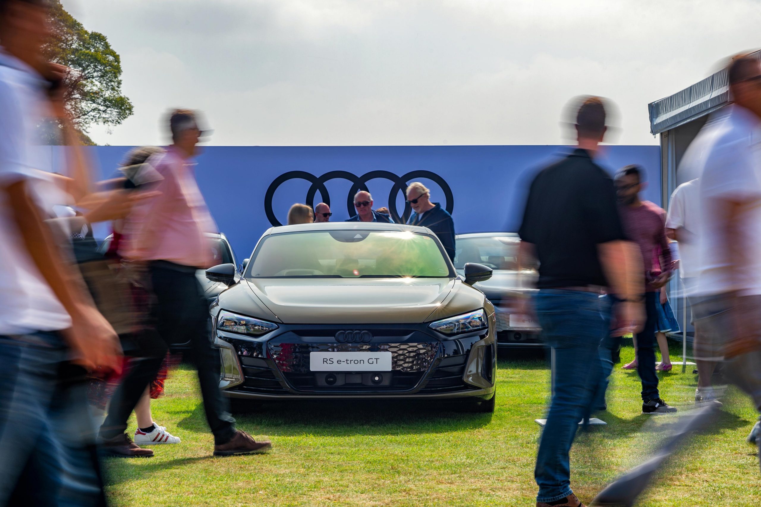 A slow shutter speed image captures the bustling movement of people at a car/automotive event in London. The focus is on a black Audi sports car displayed on an Audi stand, with the Audi logo prominently visible in the background.
