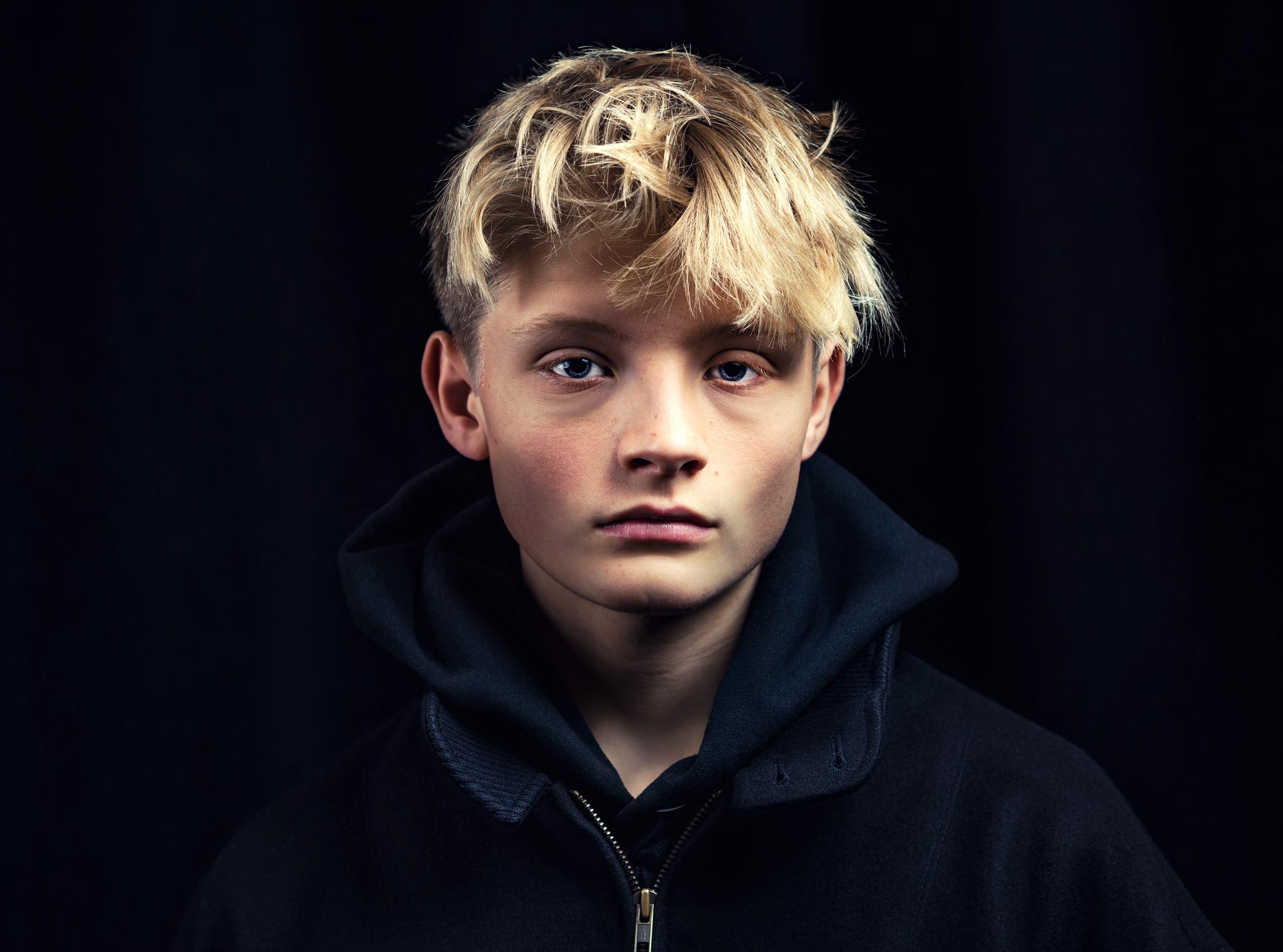 A boy model poses with a deadpan expression in a studio setting with professional lighting and a black backdrop, captured by Wright Content, a beauty photographer in London.