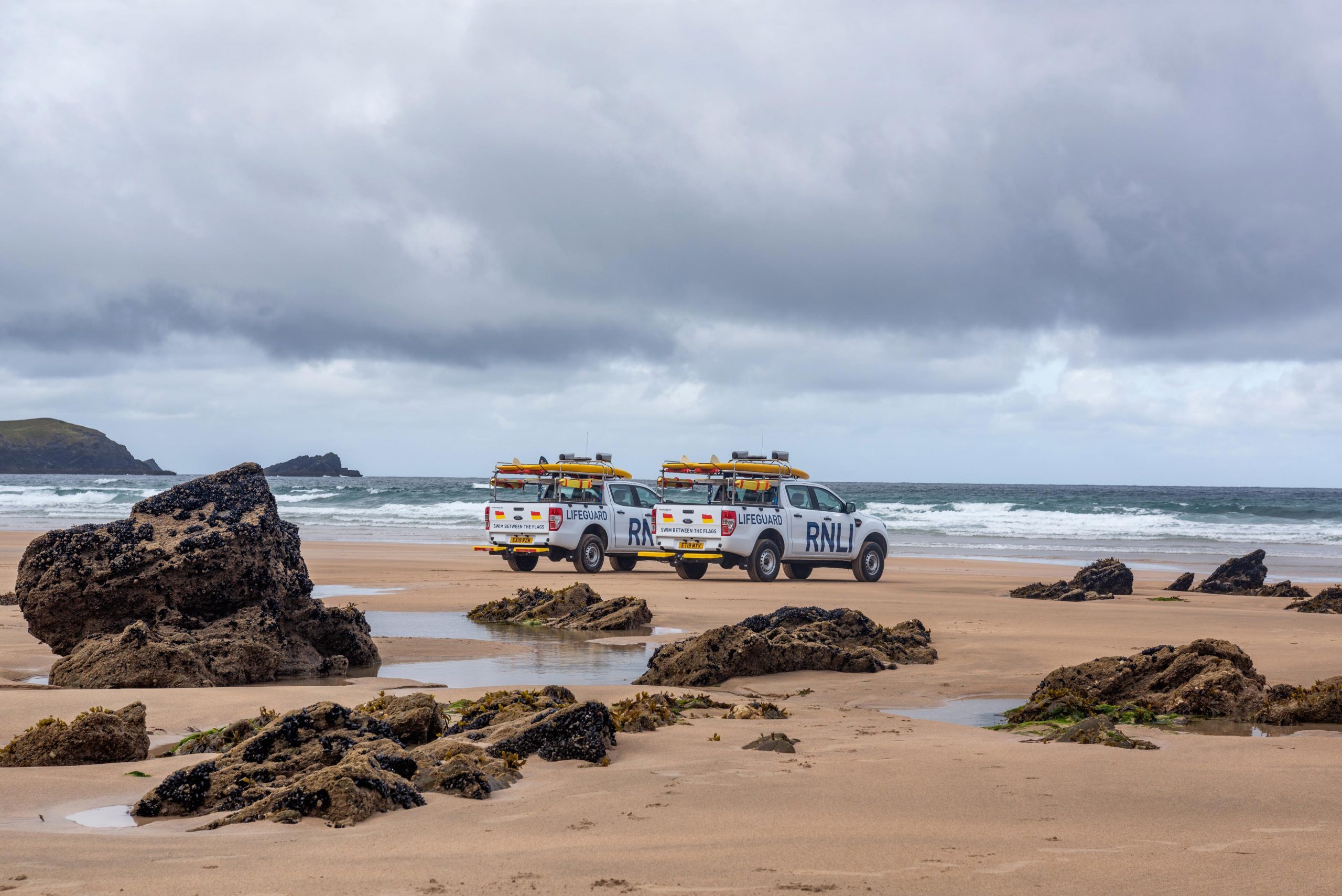 A powerful scene captured by Wright Content, featuring two Ford pickups adorned with the RNLI lifeguard logo, positioned on a beach between rugged rocks, facing the swelling sea. With surfboards mounted on top, the vehicles stand ready for action, manned by vigilant lifeguards prepared for any situation. Wright Content specializes in evocative brand photography.