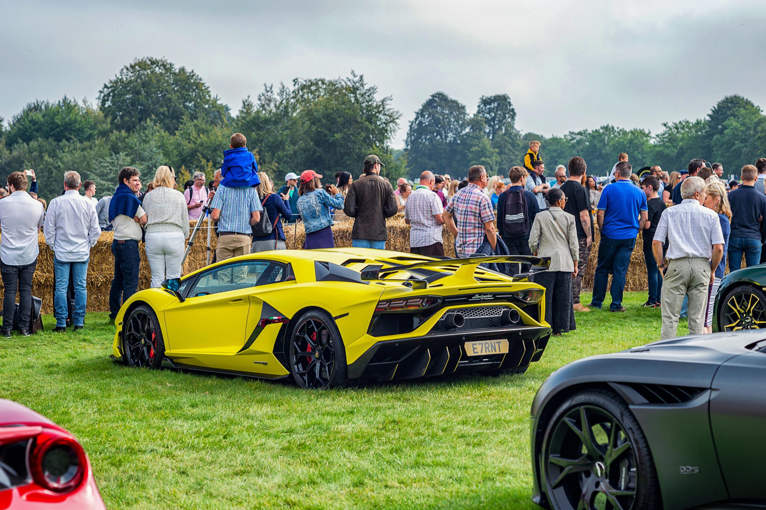 A vibrant green Lamborghini sits prominently on the grass beside a race track, with spectators lining the track, watching cars race by at a thrilling car event in London. The focus of the shot is on the striking green Lamborghini in the foreground.