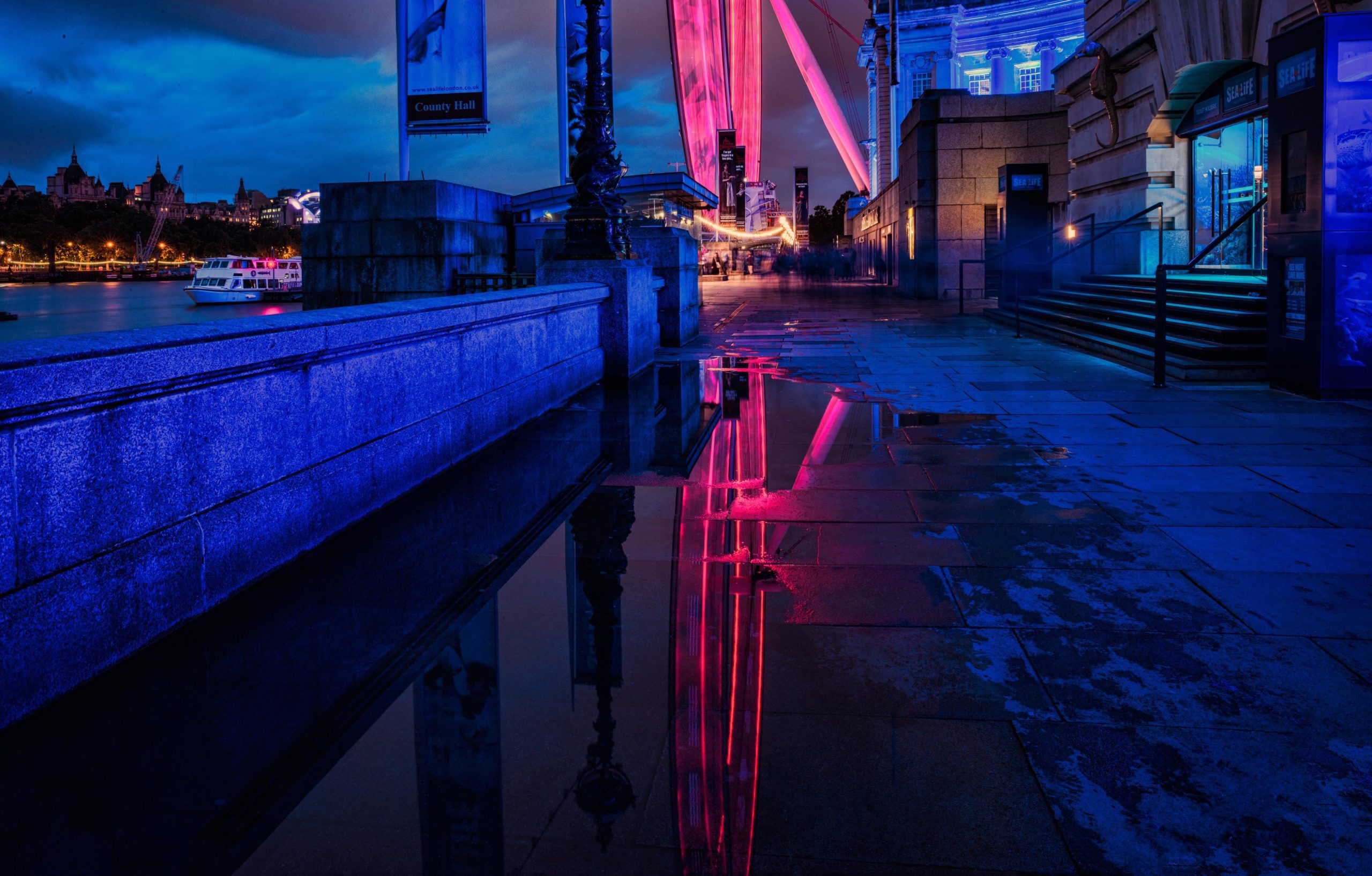 Captivating night shot of the London Eye captured by a commercial photographer in London. The long exposure image, taken from a low angle, showcases the iconic wheel in motion, creating a mesmerizing blur effect. Reflecting in a puddle, the illuminated wheel adds a sense of drama and uniqueness to the shot, perfect for the 'Epic London' project by a marketing company.