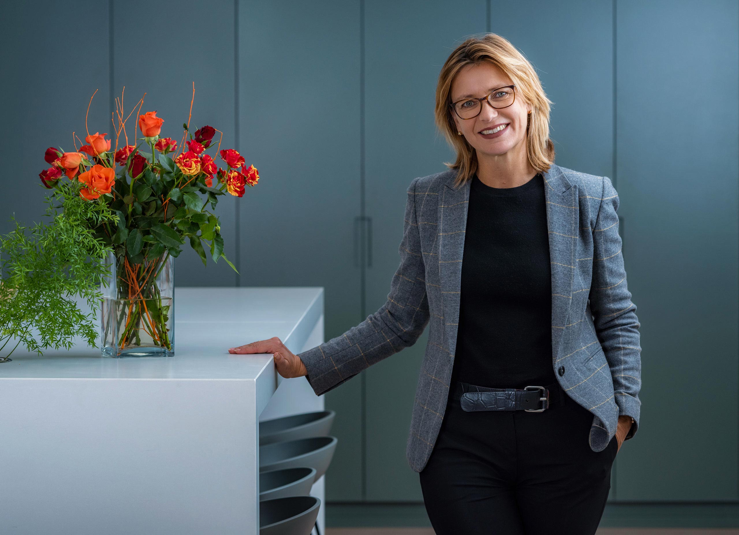 A woman managing director of a large real estate communications company in London (ING Media) stands in her office wearing a smart jacket and glasses. There are red roses in a vase on the breakfast bar next to her.