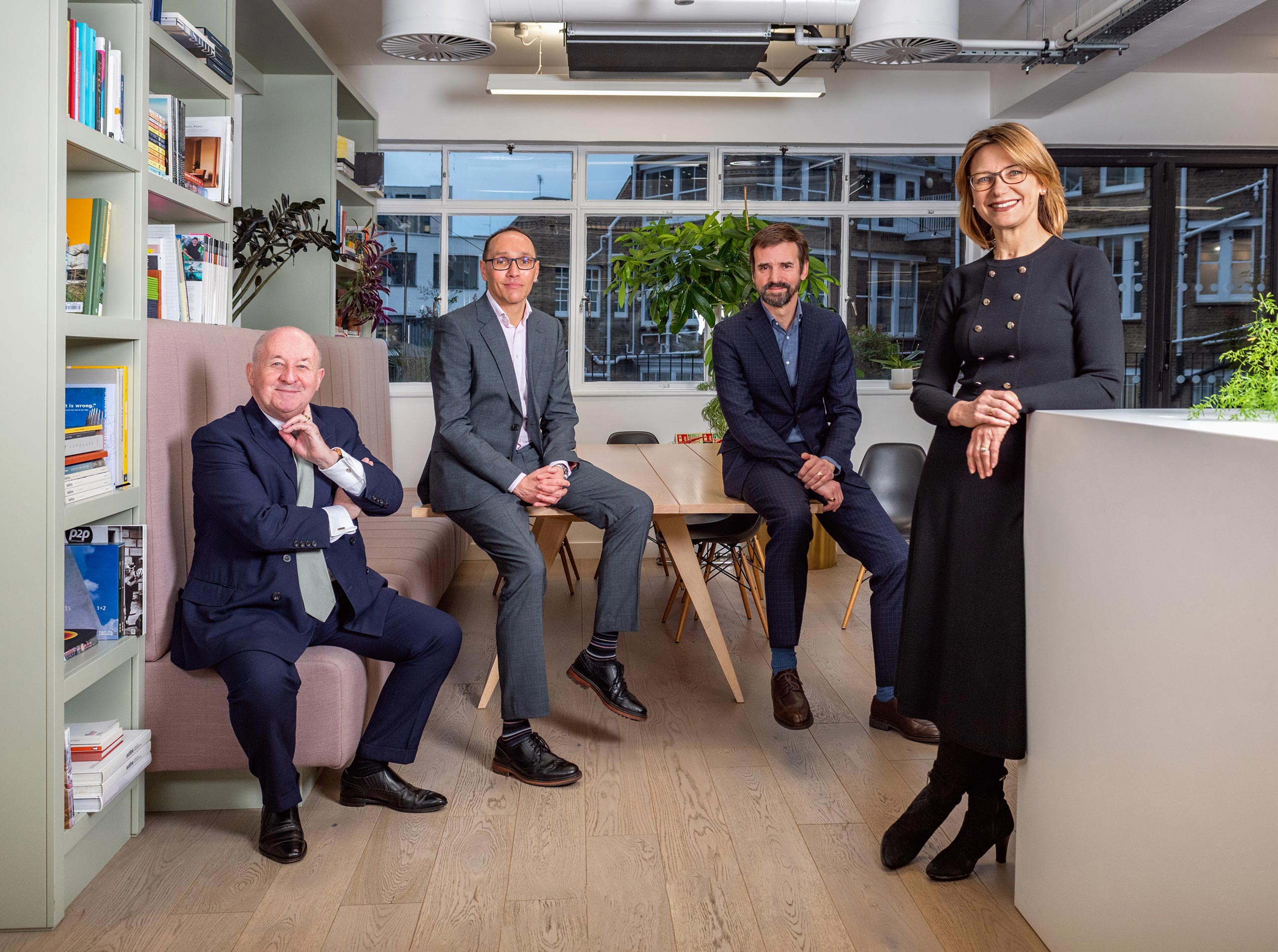 A group shot of four senior professionals standing and sitting in their London offices, casual yet formal.