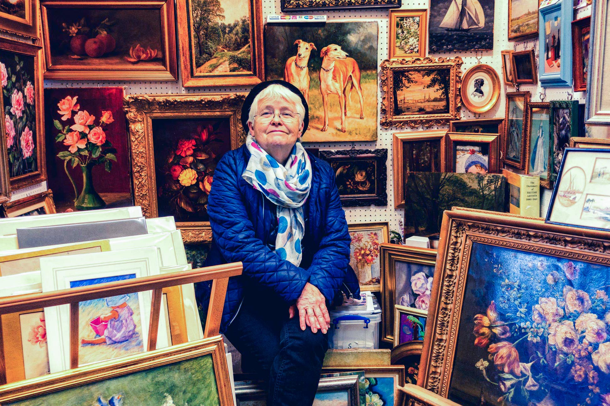 An elderly woman sells artwork in a renowned London market, surrounded by paintings in gold guild frames. Captured by Wright Content, an environmental portrait photographer in London.