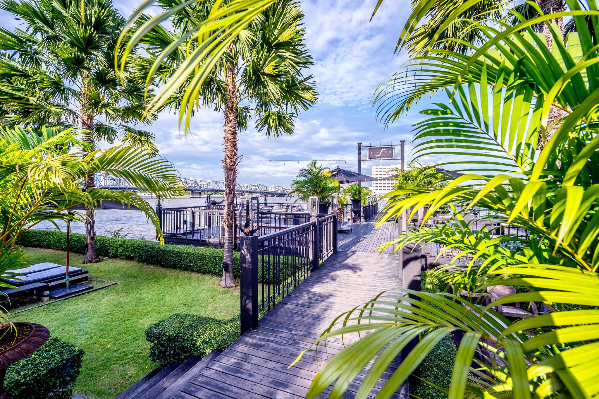 Exterior photograph of the jetty at The Siam, a luxurious hotel in Bangkok, Thailand, captured by Wright Content. The image showcases the elegant jetty surrounded by palm trees, overlooking the river with the iconic metal railing bridge in the background.