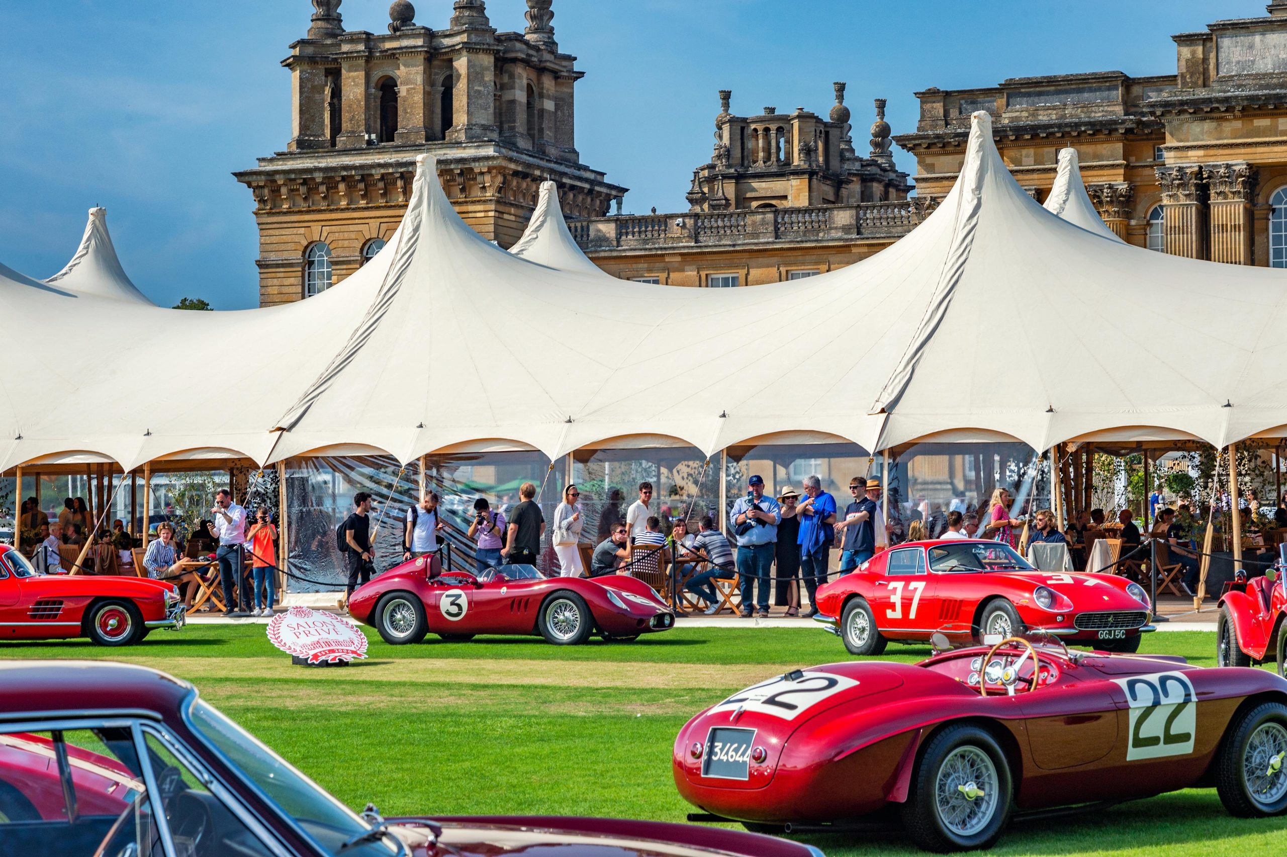 A shot of a car show event in London, featuring people enjoying the show with red classic Ferraris in the foreground and a large Manor House in the background.