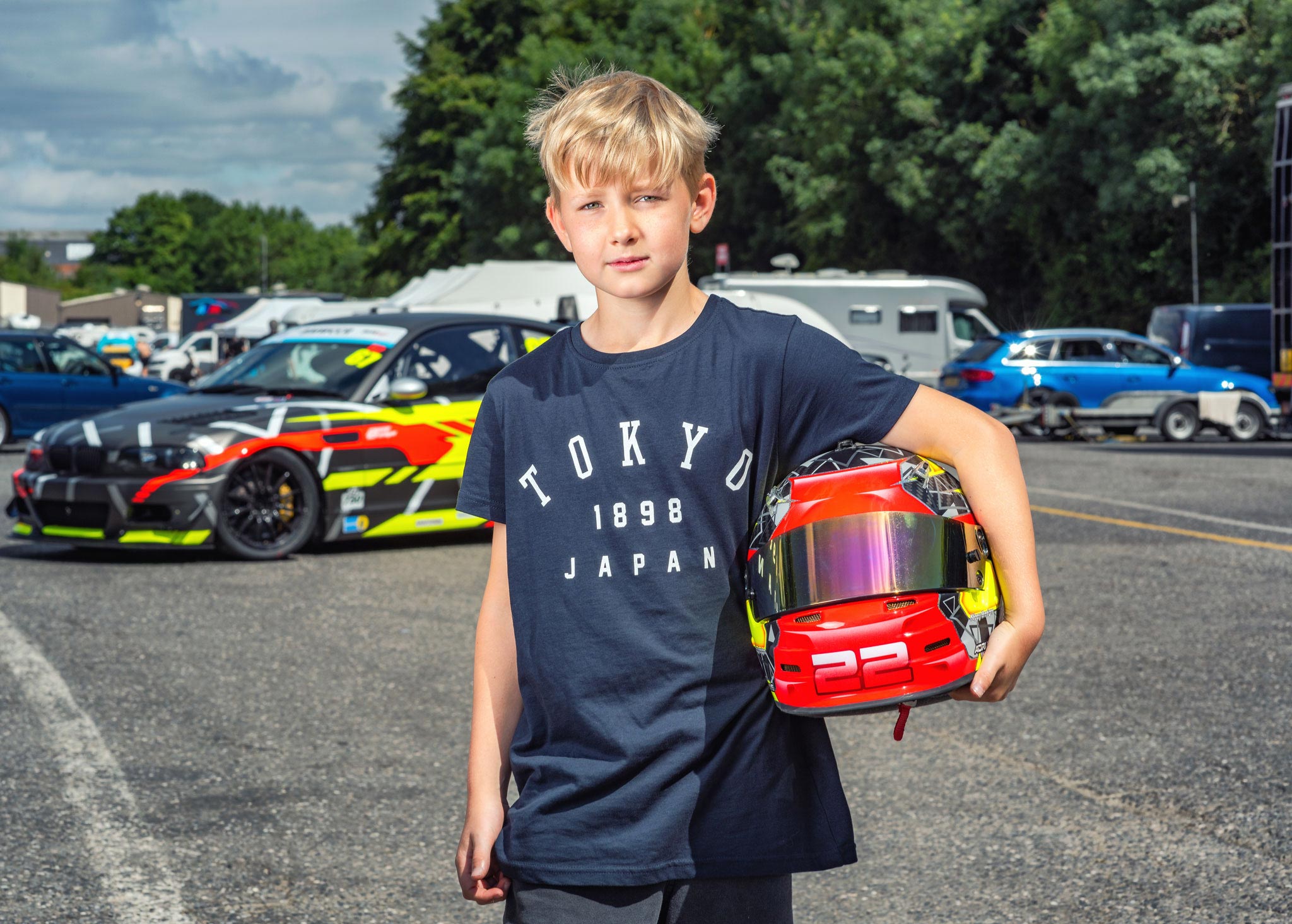 A boy poses for his portrait holding a race car driver's helmet during a track day event in motorsport. Captured by Wright Content, a family portraits photographer in London.
