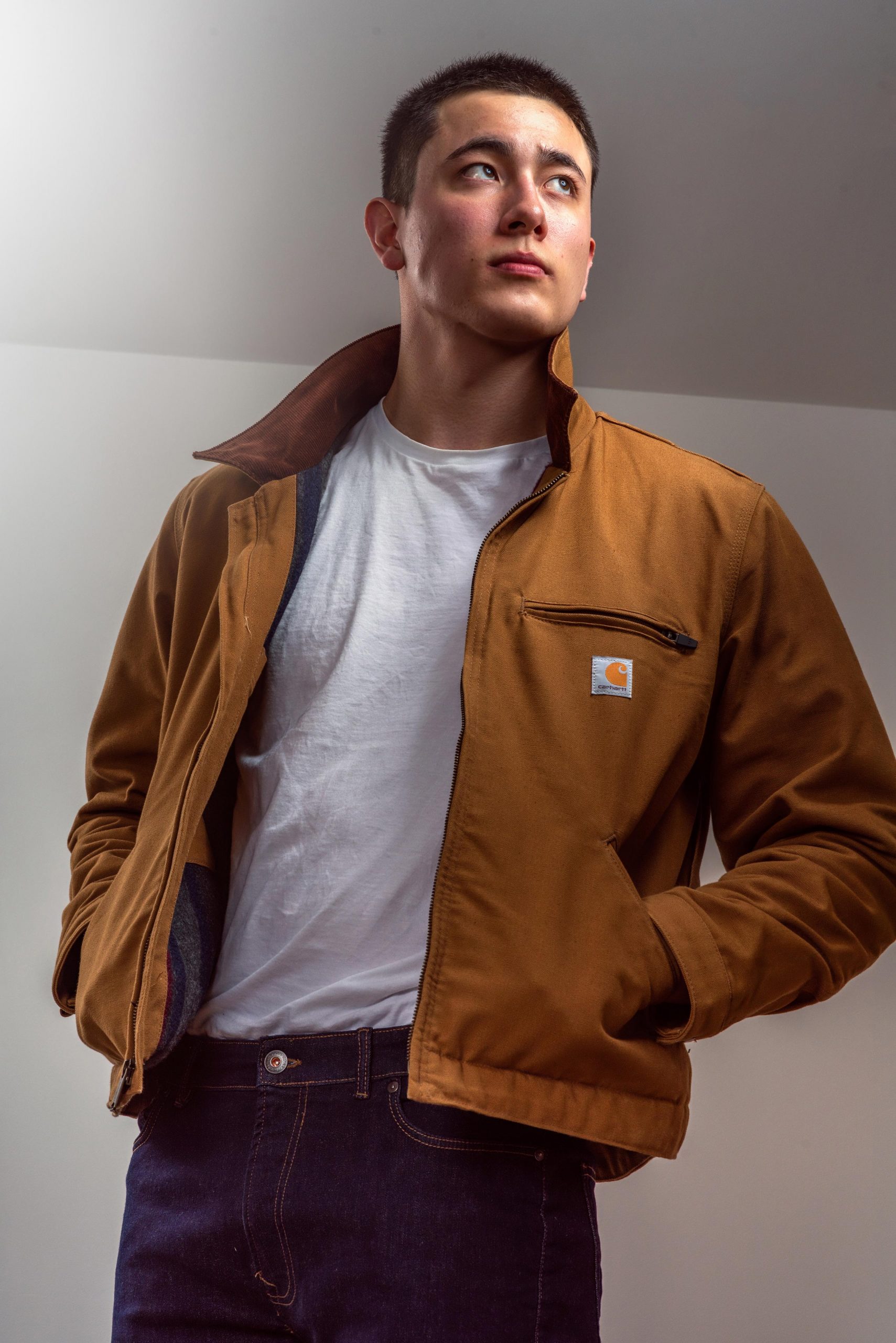 A male model wears a Carhartt jacket in a studio setting, illuminated by soft, flattering lights. He wears blue jeans and a white t-shirt while gesturing with the jacket, hands in the sandy-colored jacket's pockets.