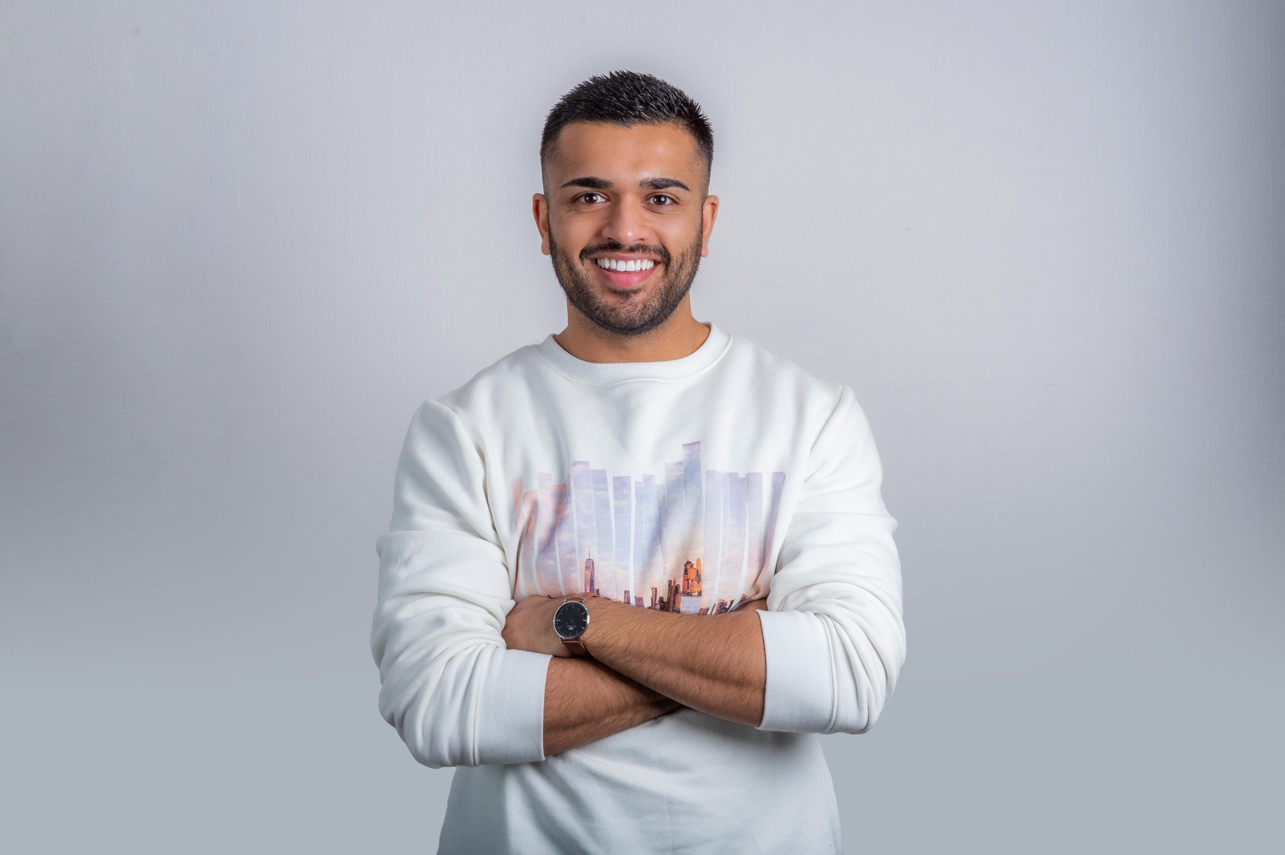 Confident young man poses with folded arms and a bright smile for headshot photography, representing a tech company, against a white background.