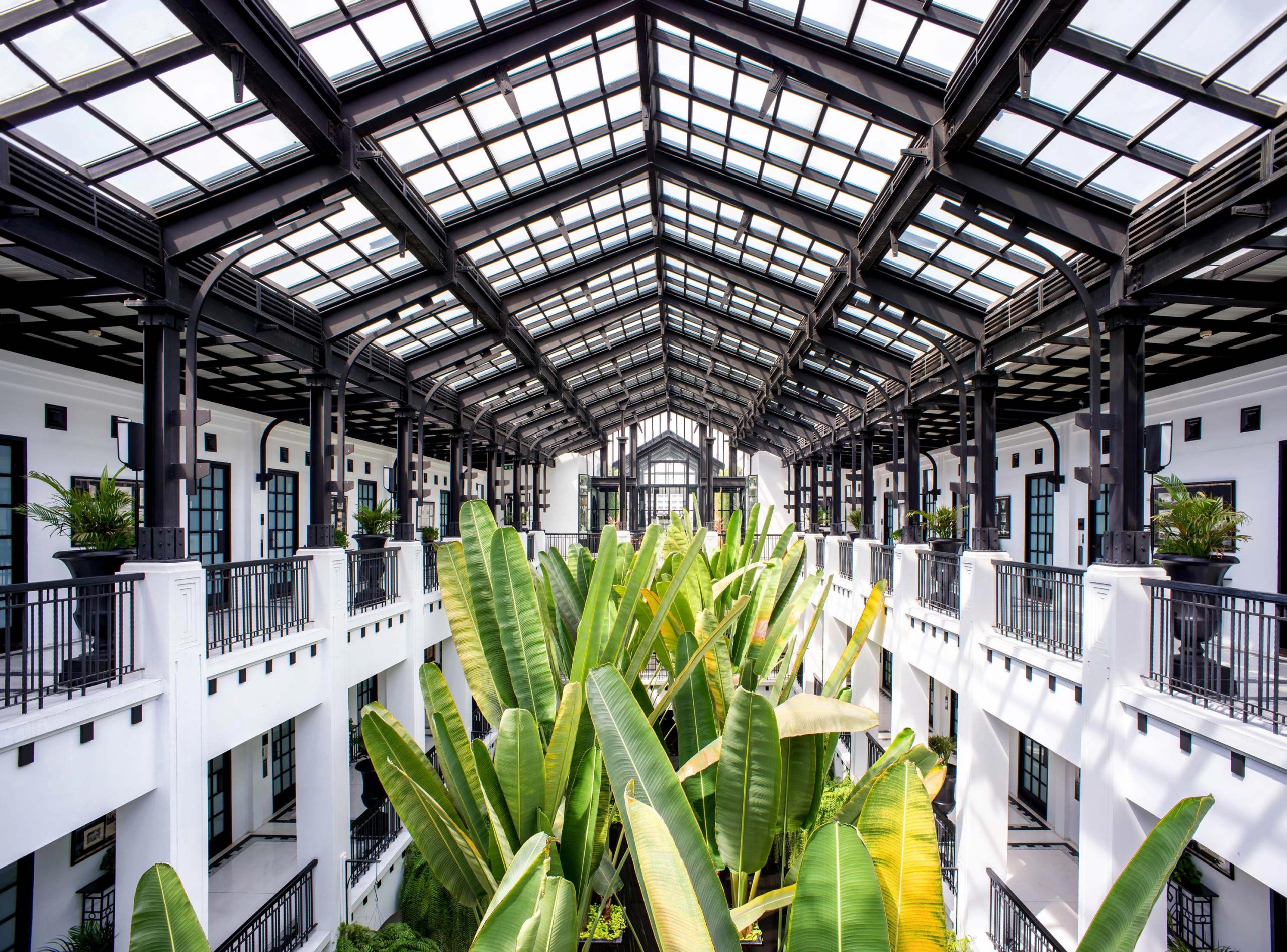Aerial view of the glass ceiling courtyard at The Siam Hotel in Bangkok, featuring tall palm trees surrounded by balconies on multiple floors.