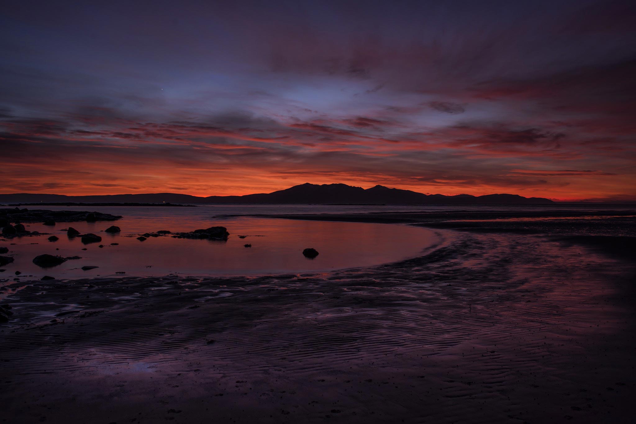 Capture the stunning beauty of West Kilbride Beach in Scotland at sunset, showcasing the ethereal glow of the sky with hues of red and blue. The landscape features the tranquil sea stretching towards the horizon, with the silhouette of Arran Island in the distance. The unique lighting casts a mesmerizing purple hue on the sand and rocks, creating an otherworldly scene.