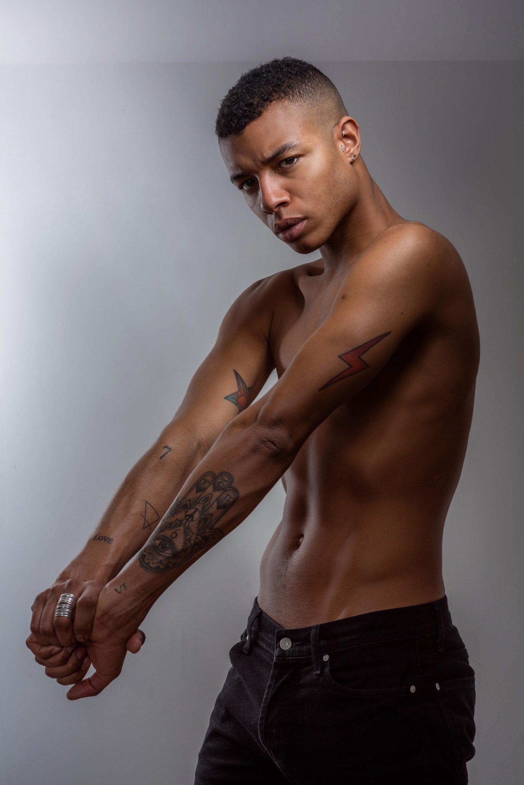A shirtless model poses in the studio with hands clasped out in front. He tilts his head and wears a serious expression, showcasing his golden brown skin adorned with tattoos.