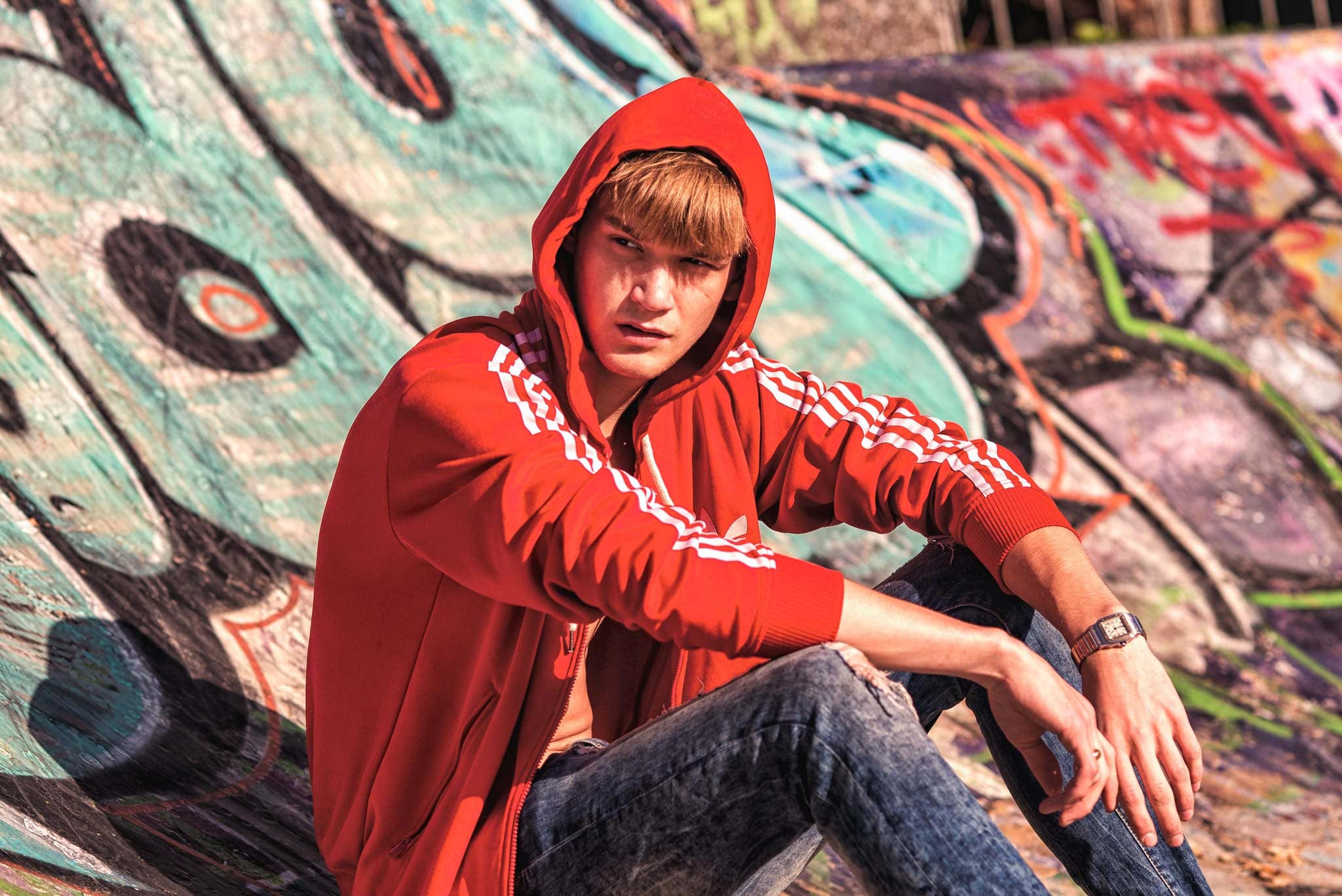 A boy fashion model sits on a skate ramp in a skate park with graffiti in the background. He wears a red tracksuit top and jeans, with the sun shining in his eyes as he looks away from the camera with an inquisitive look. Captured by Wright Content, a model photographer in London.