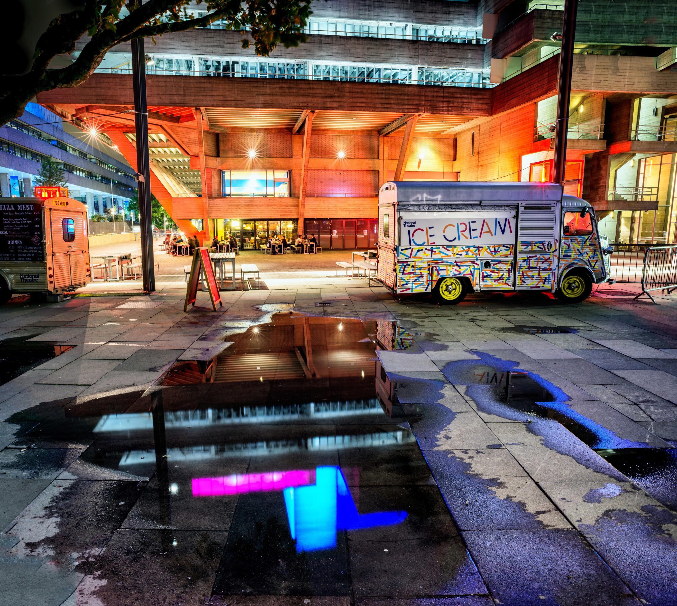 A captivating night street shot of the National Theatre exterior architecture in London, featuring vibrant neon lights reflected in puddles. An ice cream van adds a touch of urban charm to the foreground against the backdrop of the iconic brutalist architecture.