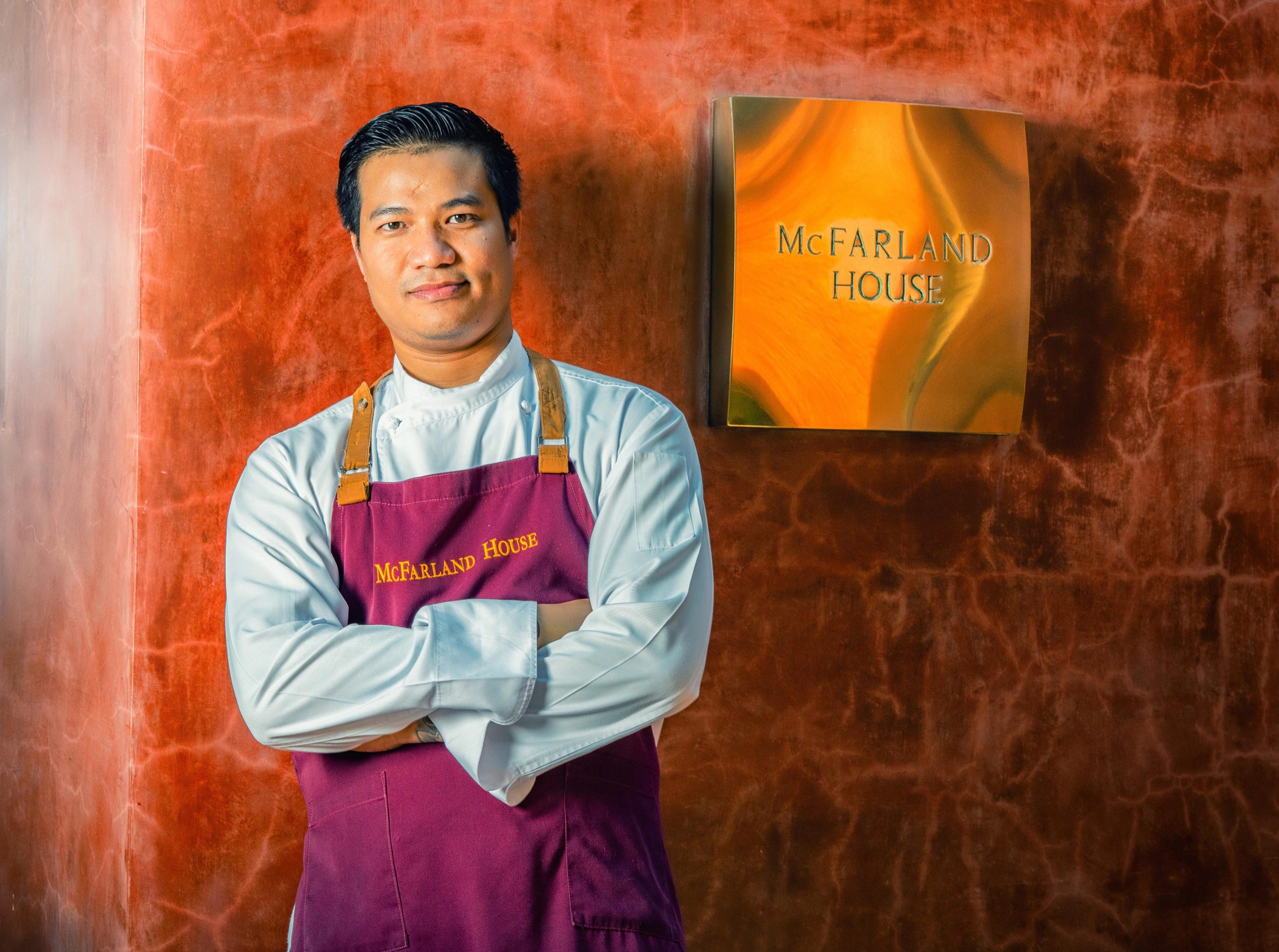 A confident chef poses outside his restaurant 'McFarland House' in the sun, shot in Thailand for a hotel chain. Captured by Wright Content, a portrait photographer.