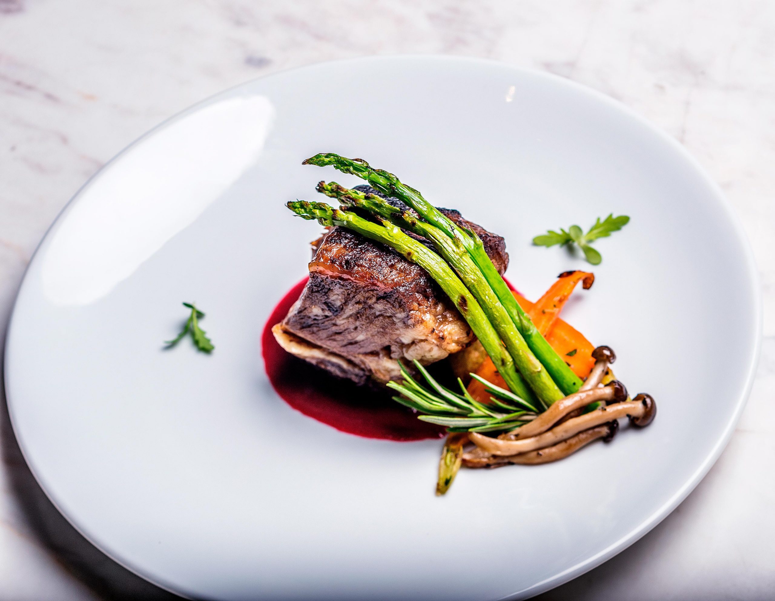 A meticulously polished food shot captures the essence of culinary excellence. The image showcases a beautifully plated dish featuring steak, asparagus, carrot, and mushrooms, set on a white plate atop a pristine white marble table. Expertly lit and crisply detailed, this food photography shot embodies the sophistication of high-end dining.