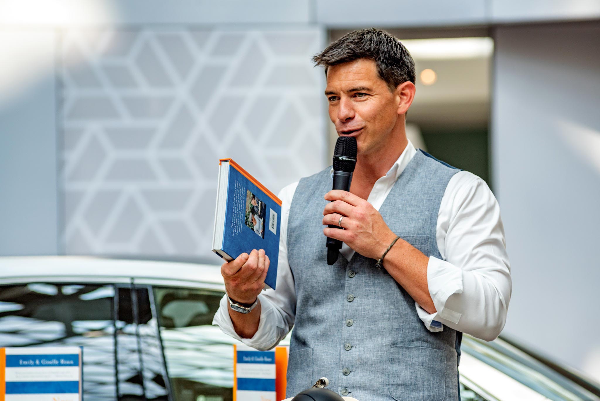 An engaging shot by Wright Content, featuring Marcus Bean, a TV presenter chef, at a Lexus experiential event. In this image, Marcus holds a microphone in one hand and a book in the other, smiling warmly as he interacts with the audience. Wright Content excels in capturing authentic moments like this.