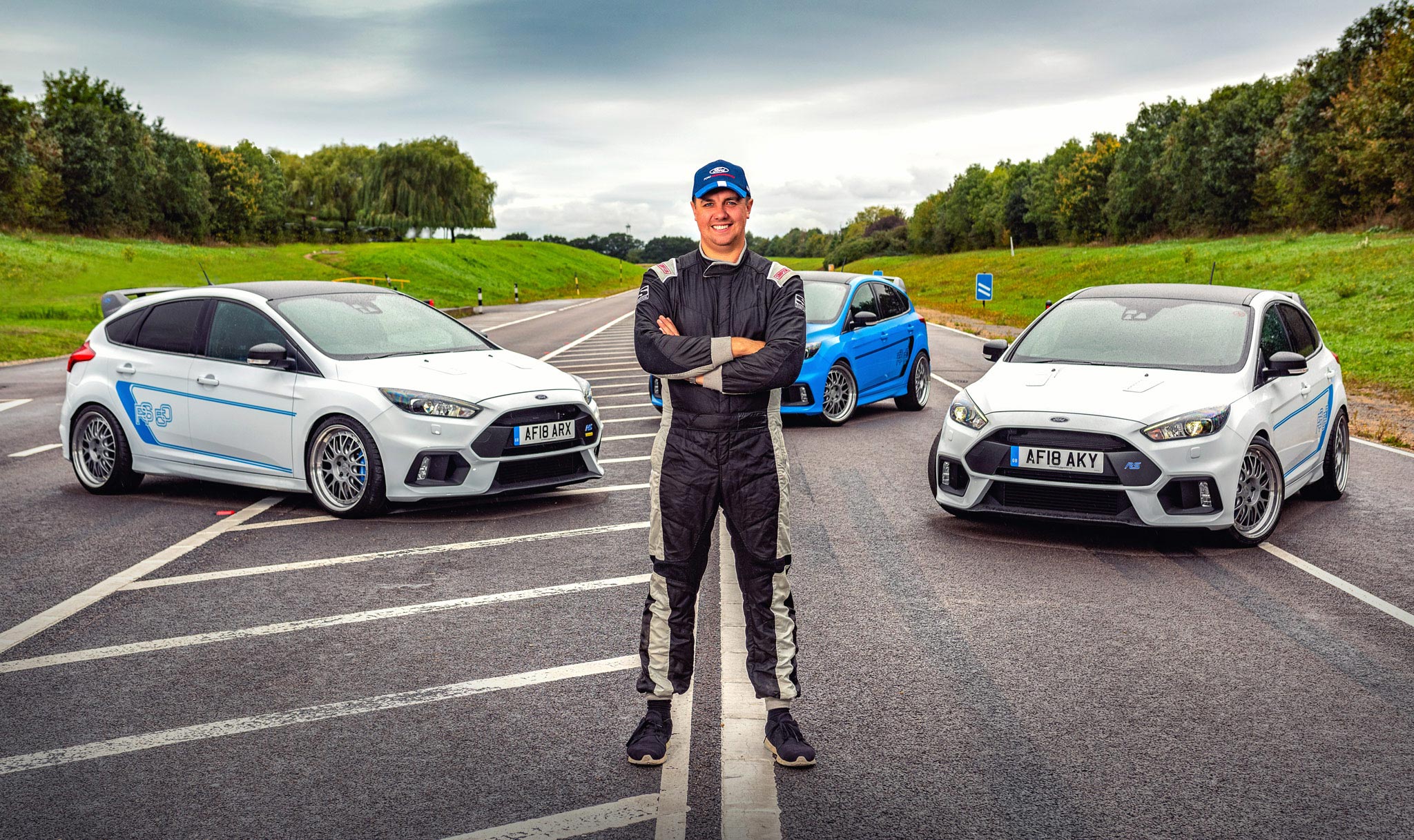 A compelling portrait captured by Wright Content, showcasing a professional driver in his racing jumpsuit and cap, standing with arms crossed against the backdrop of three Fiesta RS sports cars at Ford's test track. This image embodies the spirit of athleticism and performance, expertly documented by Wright Content, your premier sports personality photographer.