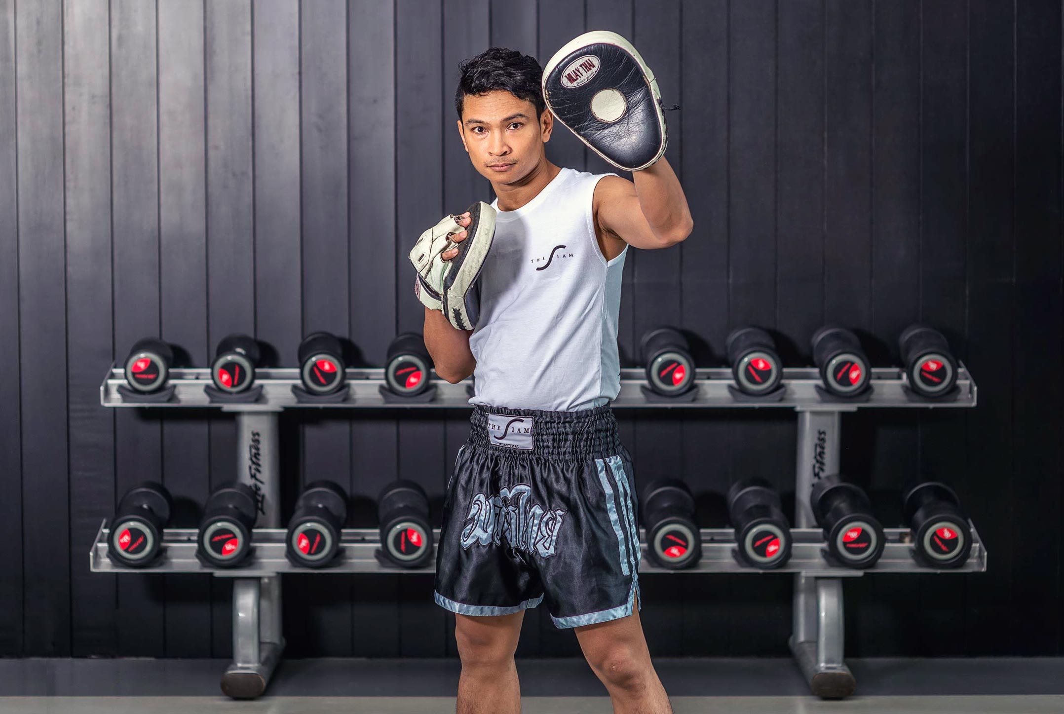 A boxing coach wearing sparring pads on his hands teaches Thai boxing for a hotel brand in Bangkok, Thailand. In the background, there is a set of dumbbells. Captured by Wright Content, a sports photographer in London.