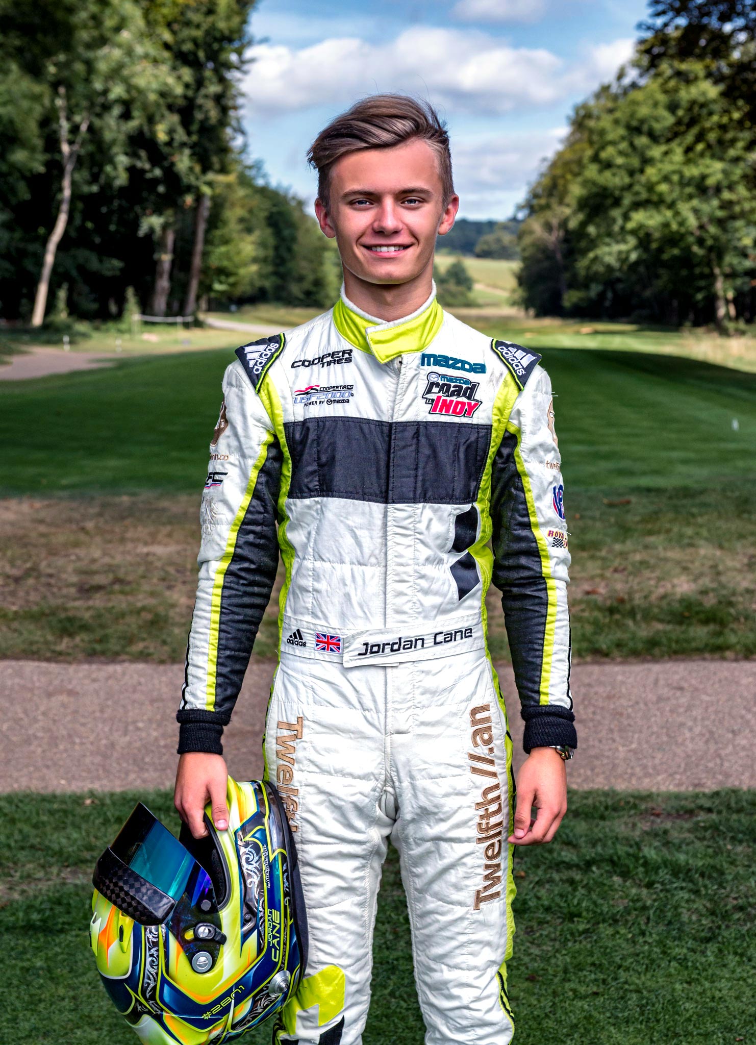 A young race car driver poses for a sports personality portrait, wearing his driving jumpsuit and holding his helmet in one hand, photographed by Wright Content, a sports portrait photographer.