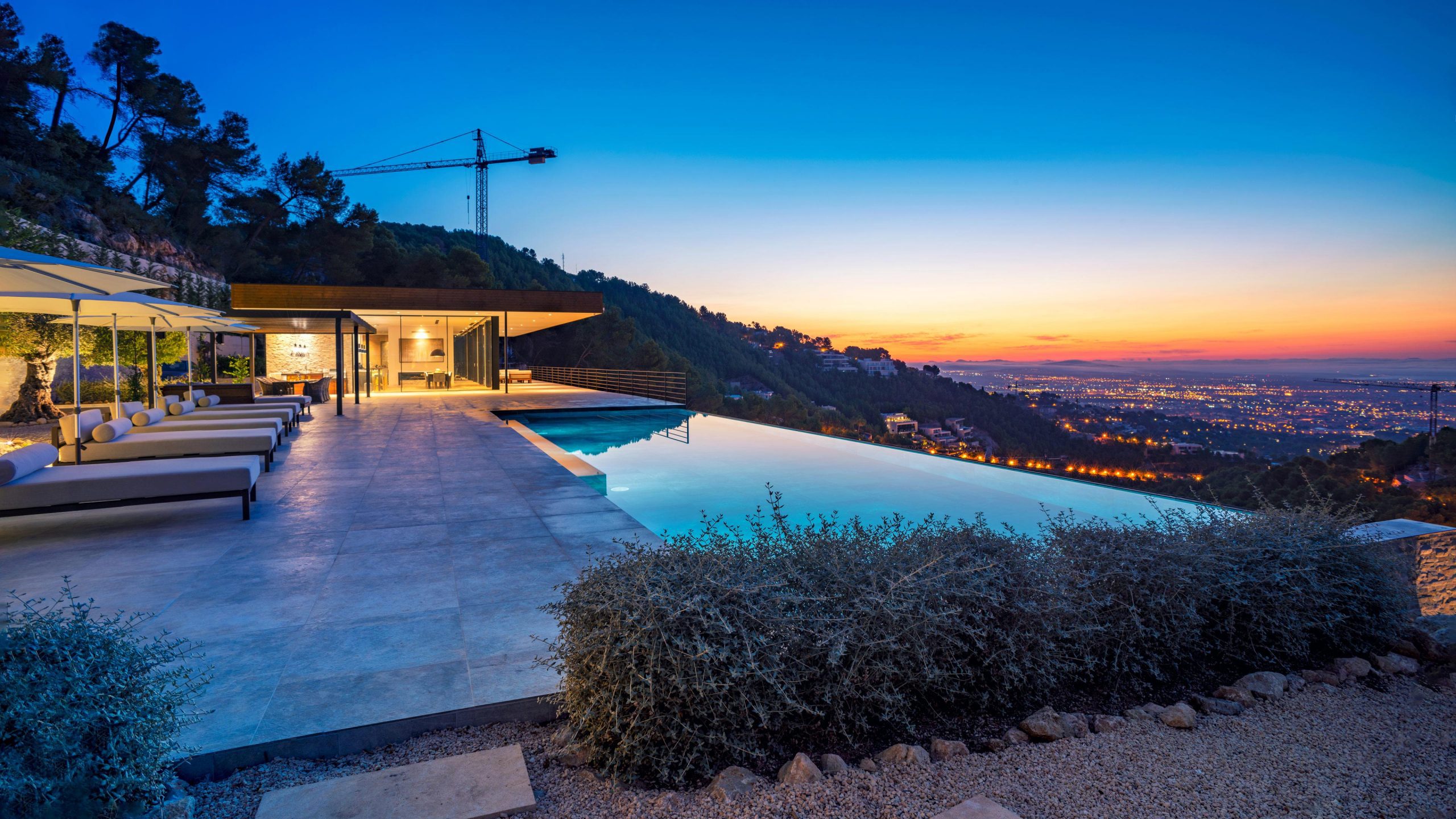 An enchanting infinity pool overlooking the city of Palma, captured at sunset by Wright Content. The city lights twinkle in the background as the poolside sun loungers invite relaxation in this cliff-top modernist home.