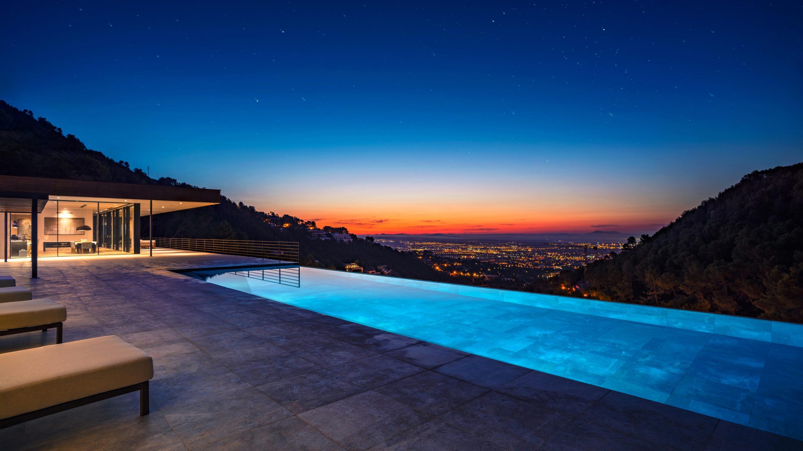 Twilight view captured by swimming pool photographer in Palma, showcasing a luxury villa atop a hill with a modernist building and illuminated pool. The city of Palma twinkles in the distance as night falls.