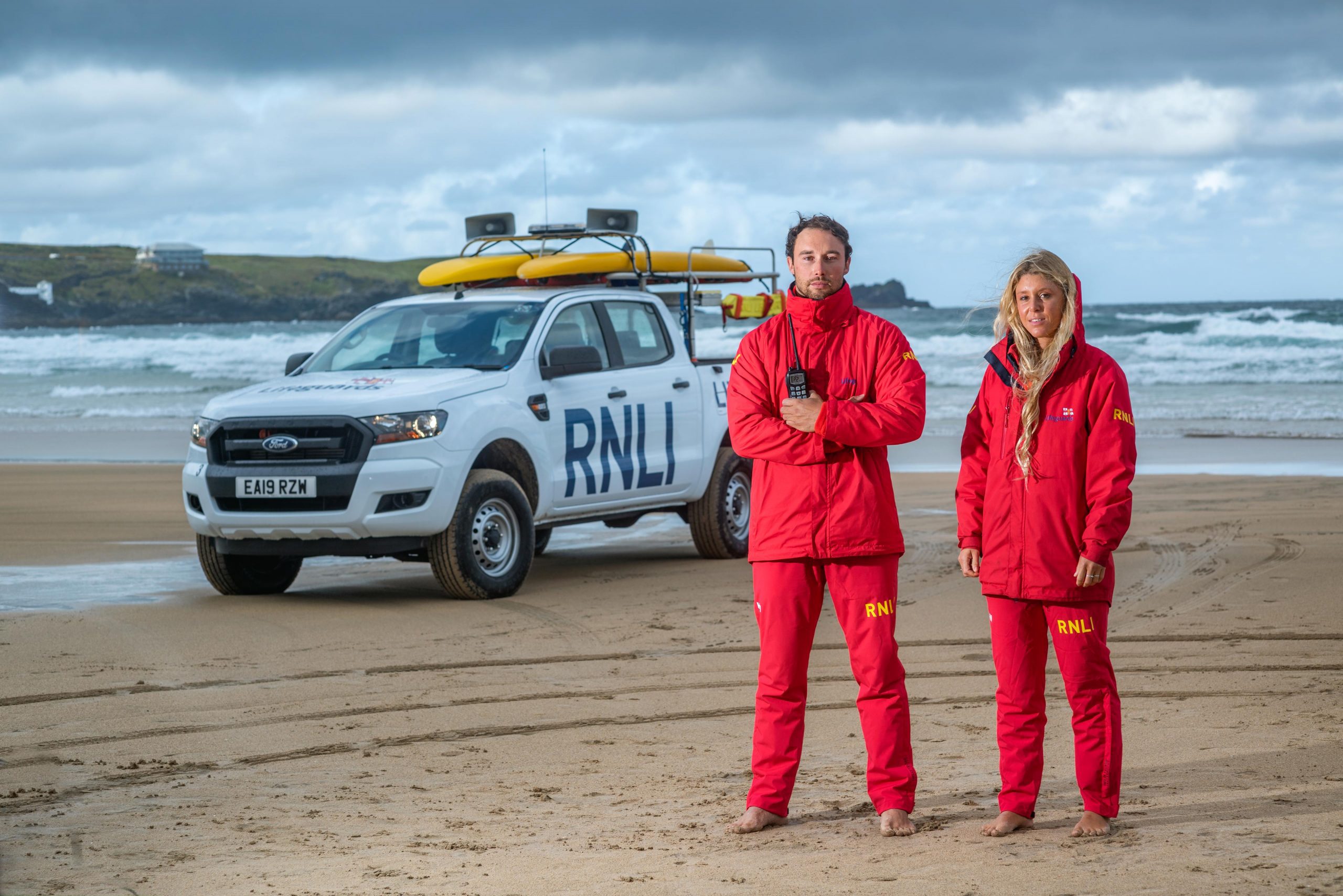 Two lifeguards from the RNLI stand on the beach in their red uniforms, with a Ford pickup truck in the background. The scene captures the dedication and readiness of the lifeguards during an open event, as captured by Wright Content." Caption: "Safety First: RNLI Lifeguards on Duty
