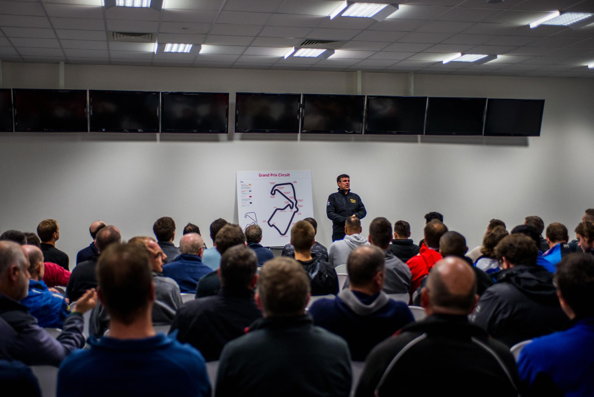 A scene captured at a corporate event briefing room, where drivers gather to prepare for an exciting day at Silverstone race track. A man stands at the front, showing a map to a large audience of drivers, briefing them about track rules and the day's race, expertly documented by Wright Content.