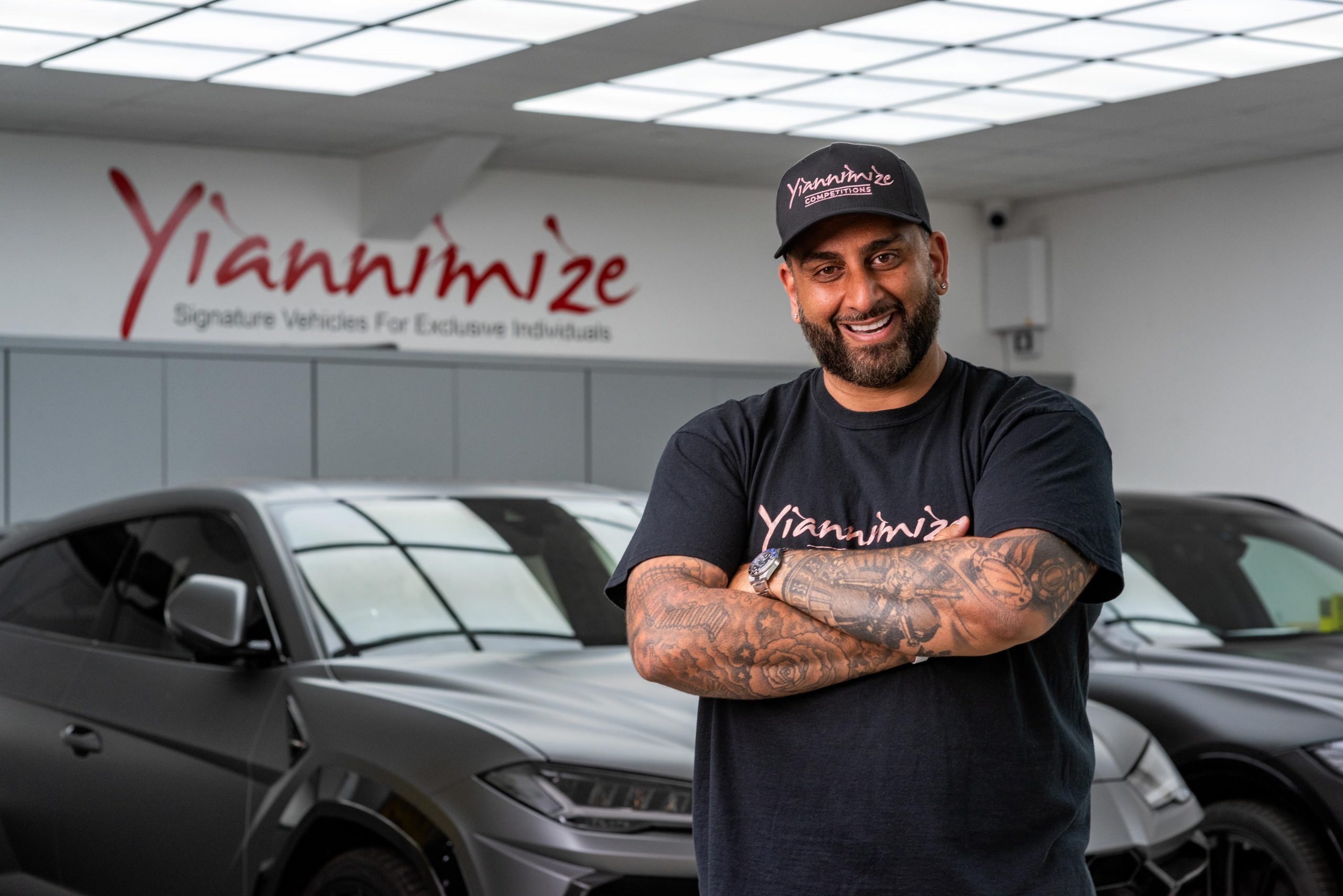 Image of social media personality Yiannimize, known for wrapping exotic cars, standing in his garage with Lamborghinis in the background. His branding is prominently displayed on the wall.