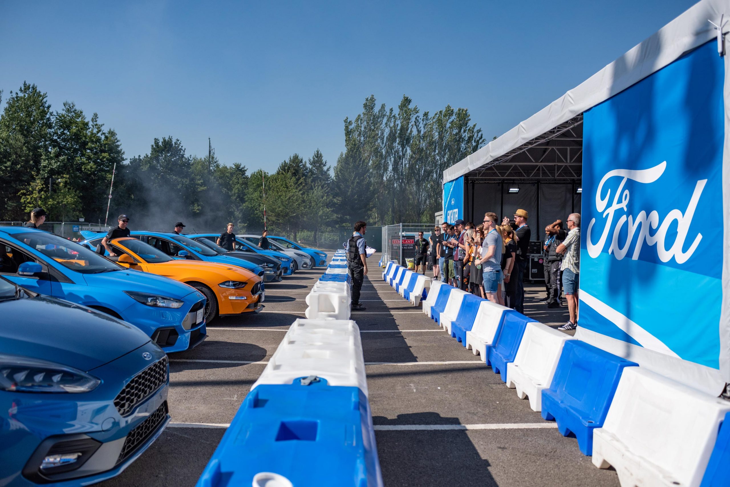 A scene of excitement captured by Wright Content, showcasing cars lined up and facing the audience at the Ford Test Centre track. Journalists invited to the event stand attentively, listening to a briefing from a professional driver in anticipation of their turn behind the wheel. Wright Content specializes in dynamic motorsport event photography.
