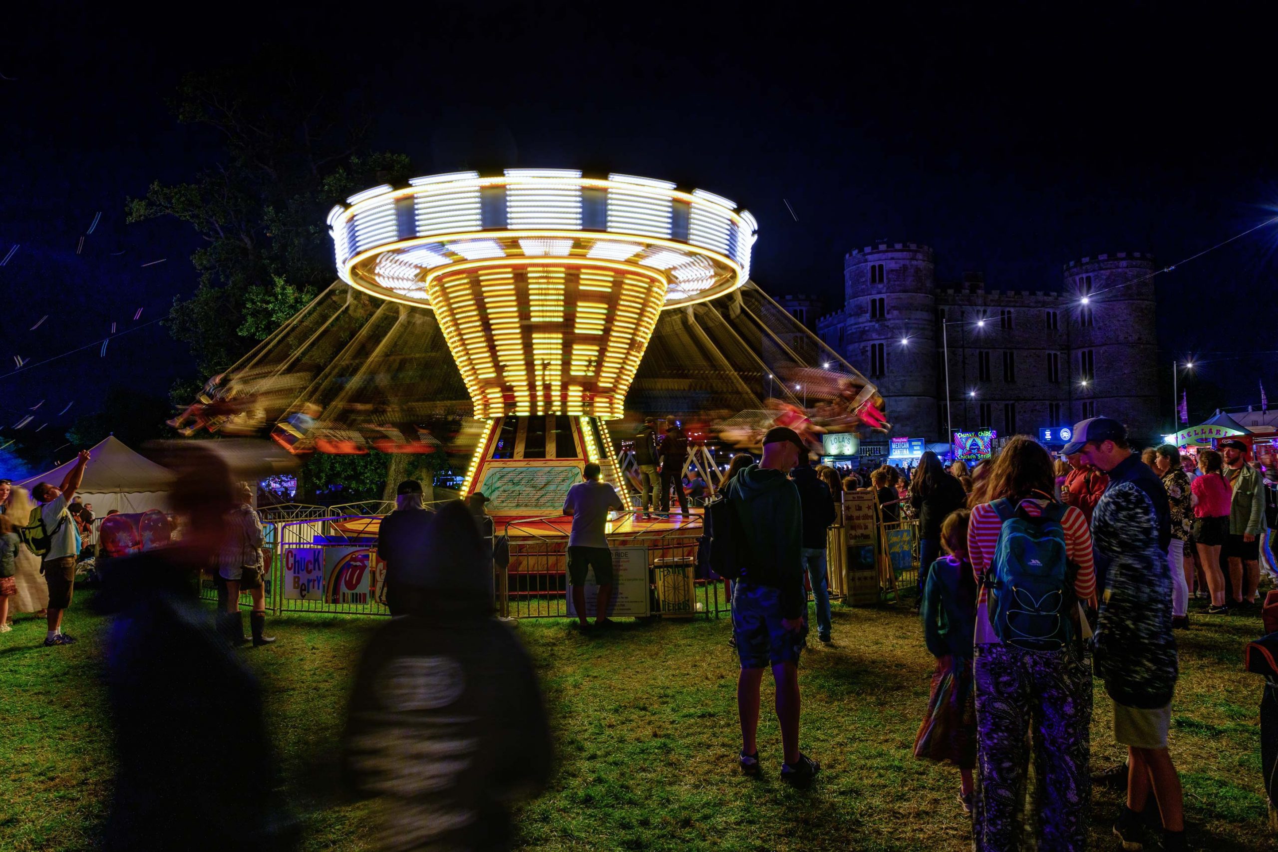 An electrifying scene captured by Wright Content at a festival, featuring party-goers enjoying the vibrant atmosphere. In the background, a fairground ride spins in the dark, illuminated by colorful lights. The slow shutter speed creates a mesmerizing effect, capturing the dynamic movement and light trails of the spinning swings. Wright Content specializes in documenting the excitement of nighttime events.